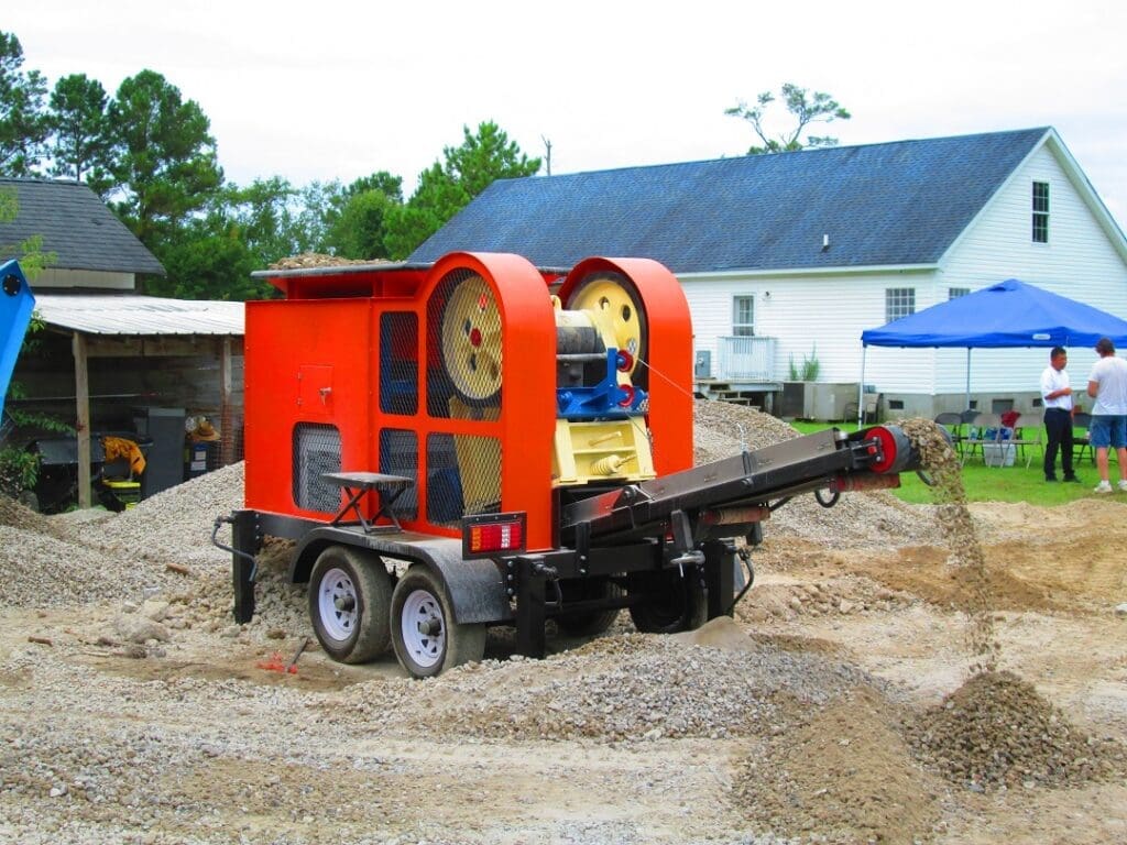 A mobile rock crusher processes gravel, discharging through a conveyor.